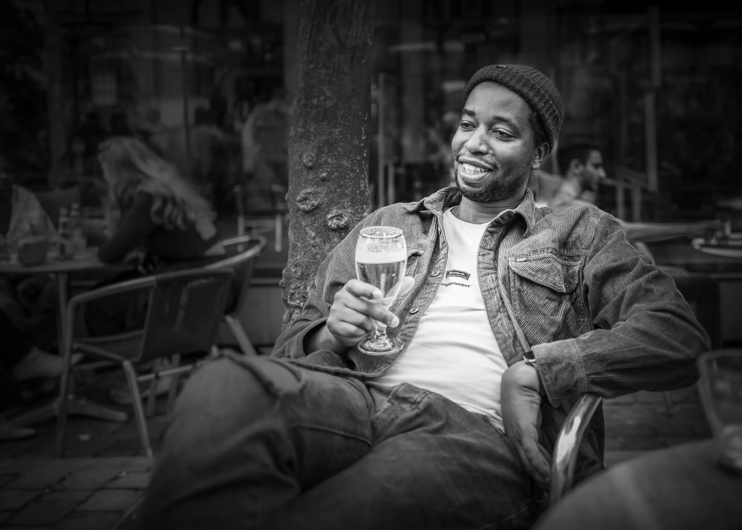 Man met muts lacht op een caféterras en houdt een glas bier vast, in zwart-wit