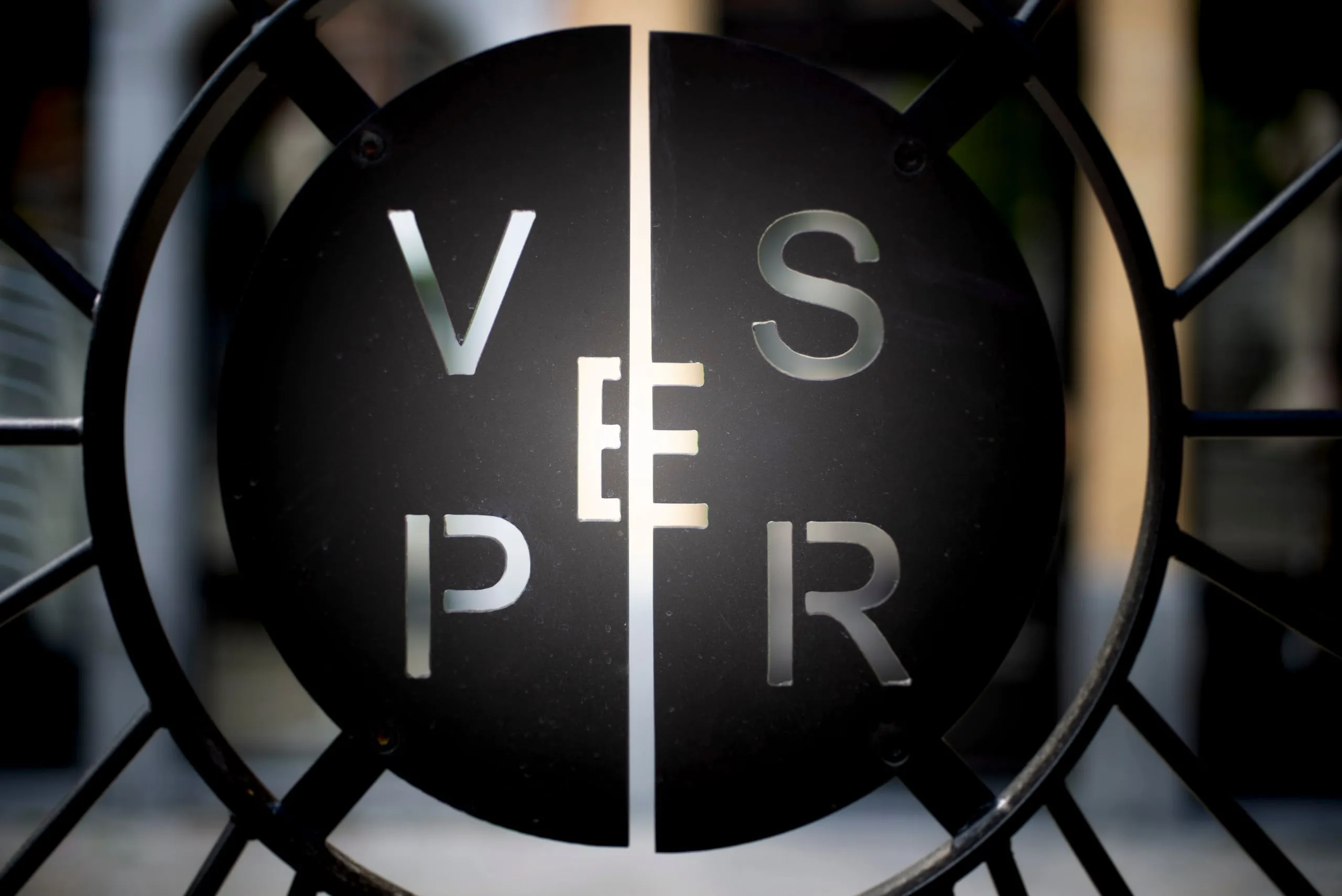 Smeedijzeren hek met een rond uithangbord met de letters van Vesper