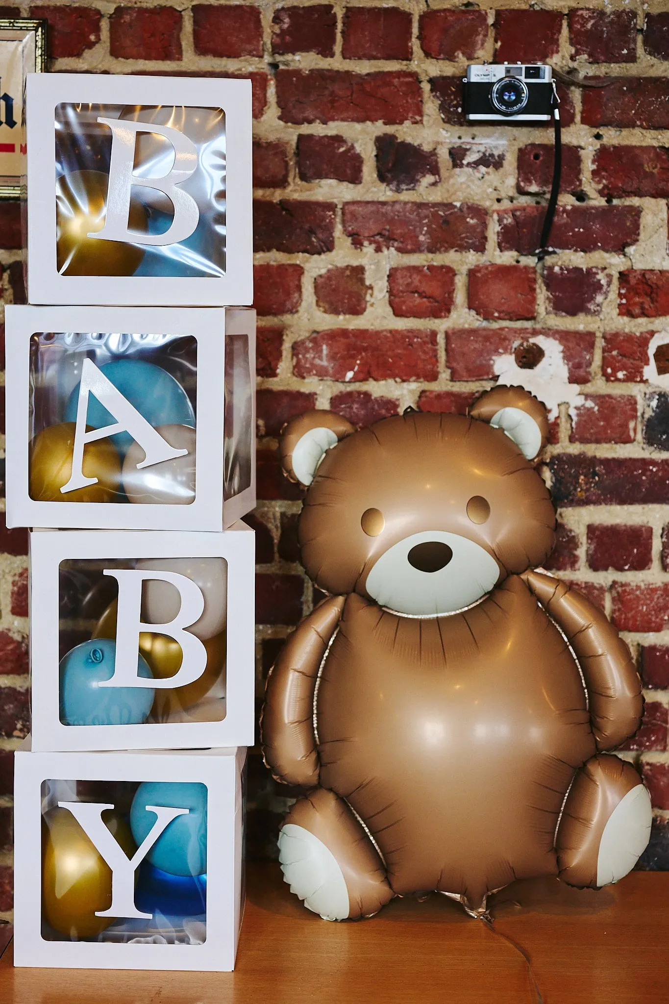 Witte letterblokken die BABY spellen naast een grote teddybeerballon tegen een bakstenen muur