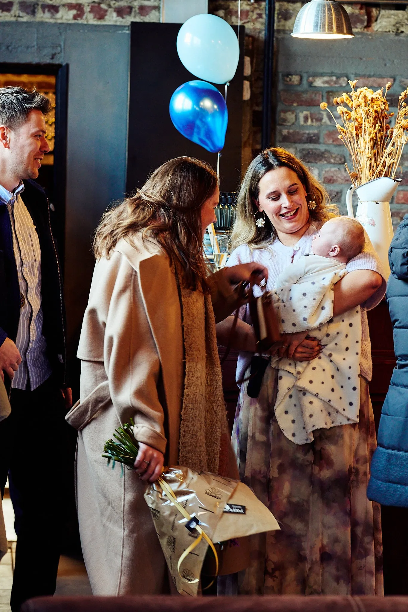 Moeder met baby in de armen begroet gasten, met ballonnen op de achtergrond