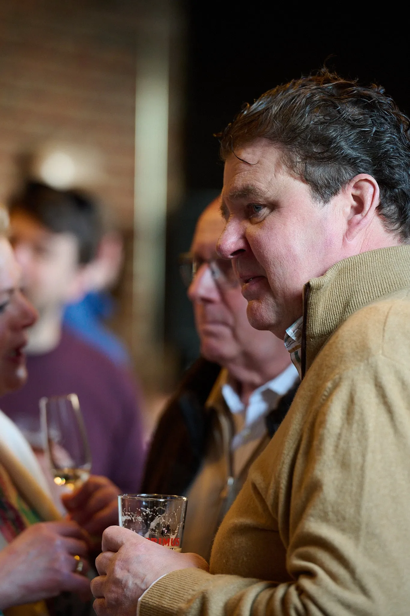 Man in beige trui met een glas in de hand tussen andere gasten