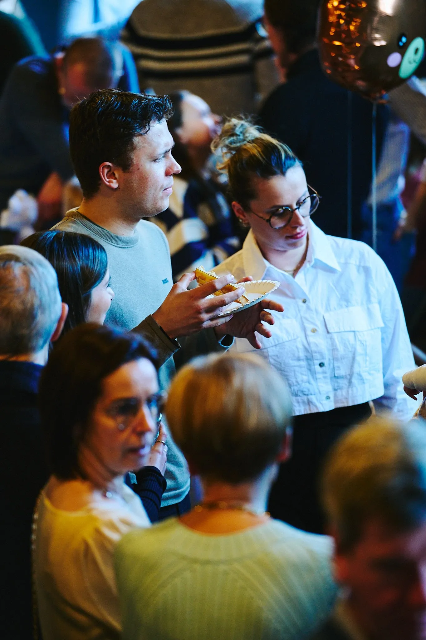 Gasten in een volle zaal, een vrouw in witte blouse houdt een bordje vast