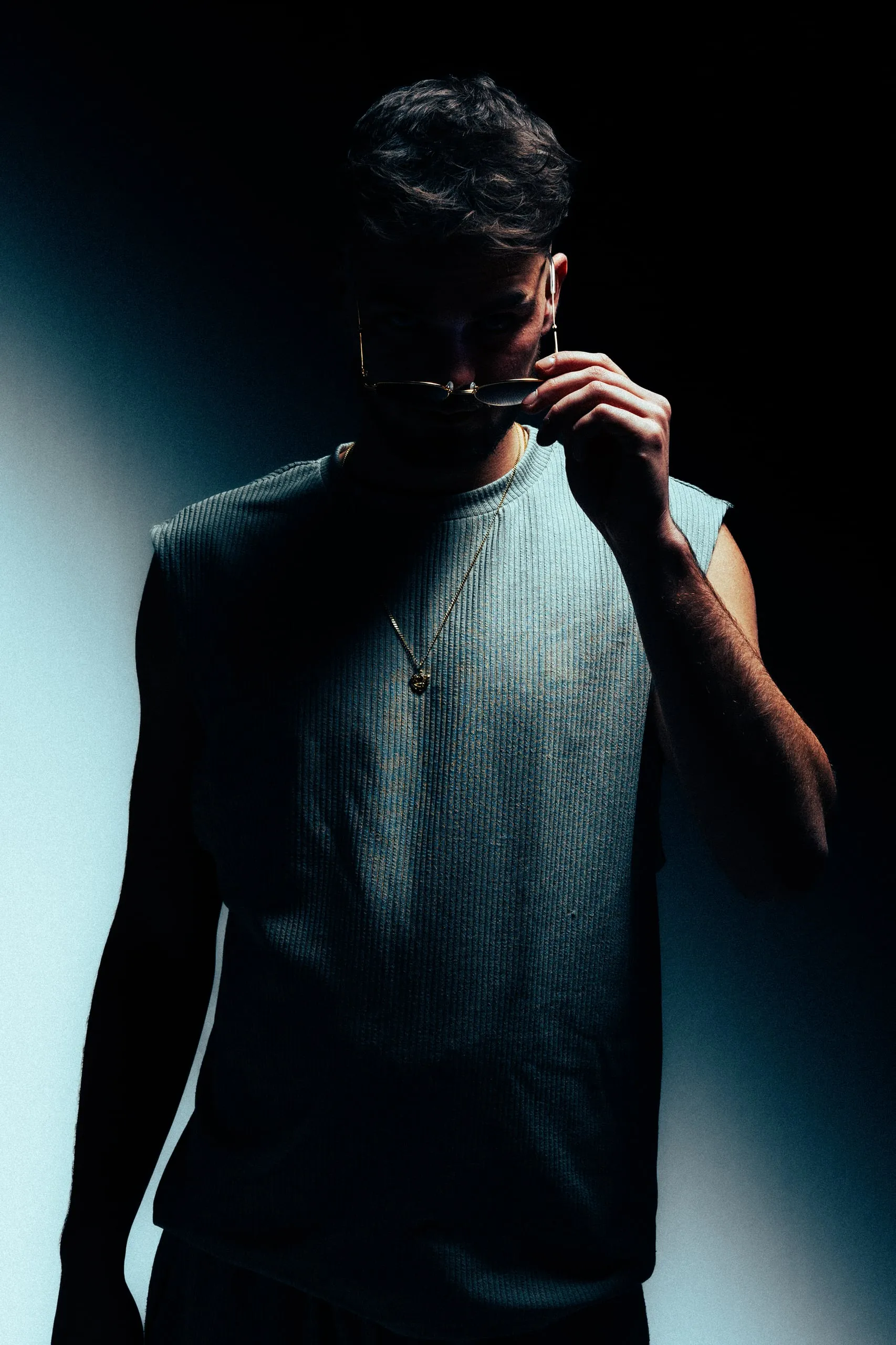 Man in mouwloos shirt houdt een bril aan zijn lippen, met scherp licht en donkere schaduwen